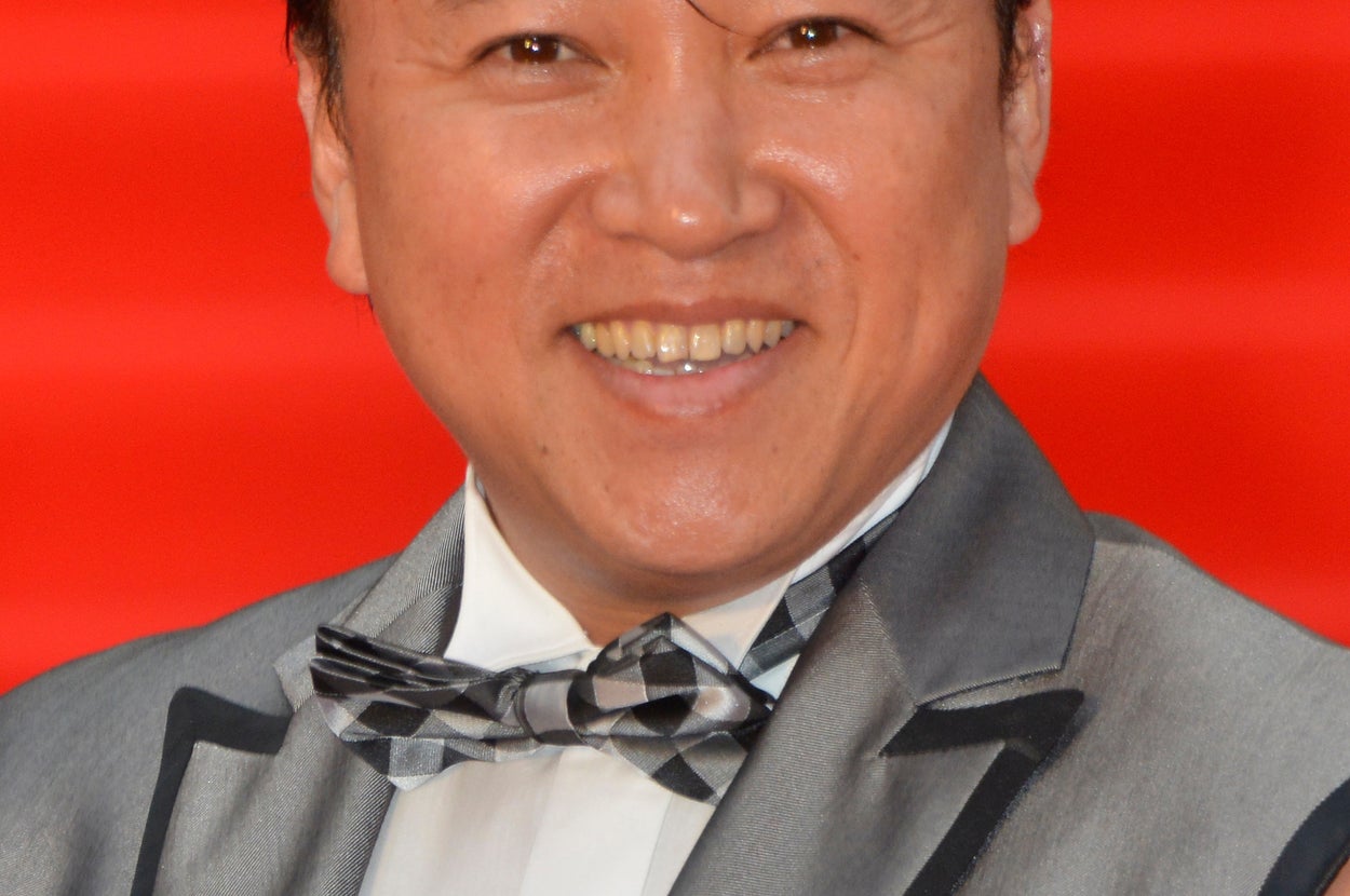 不明な人がフォーマルなスーツとユニークな蝶ネクタイを着用して、赤いカーペットで微笑んでいる様子。