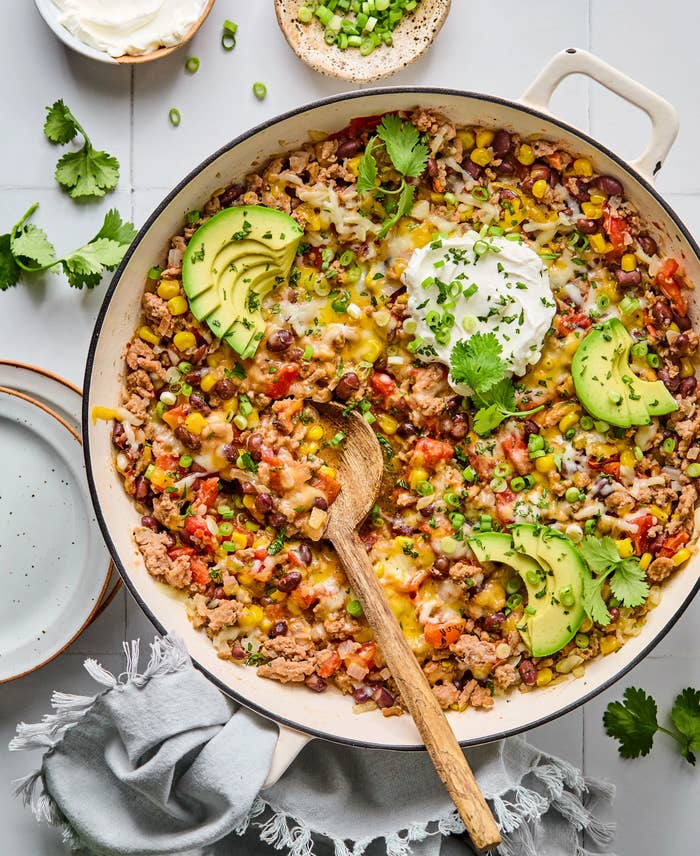 A skillet with a colorful mix of rice, beans, corn, tomatoes, cheese, and sliced avocado, topped with sour cream and herbs