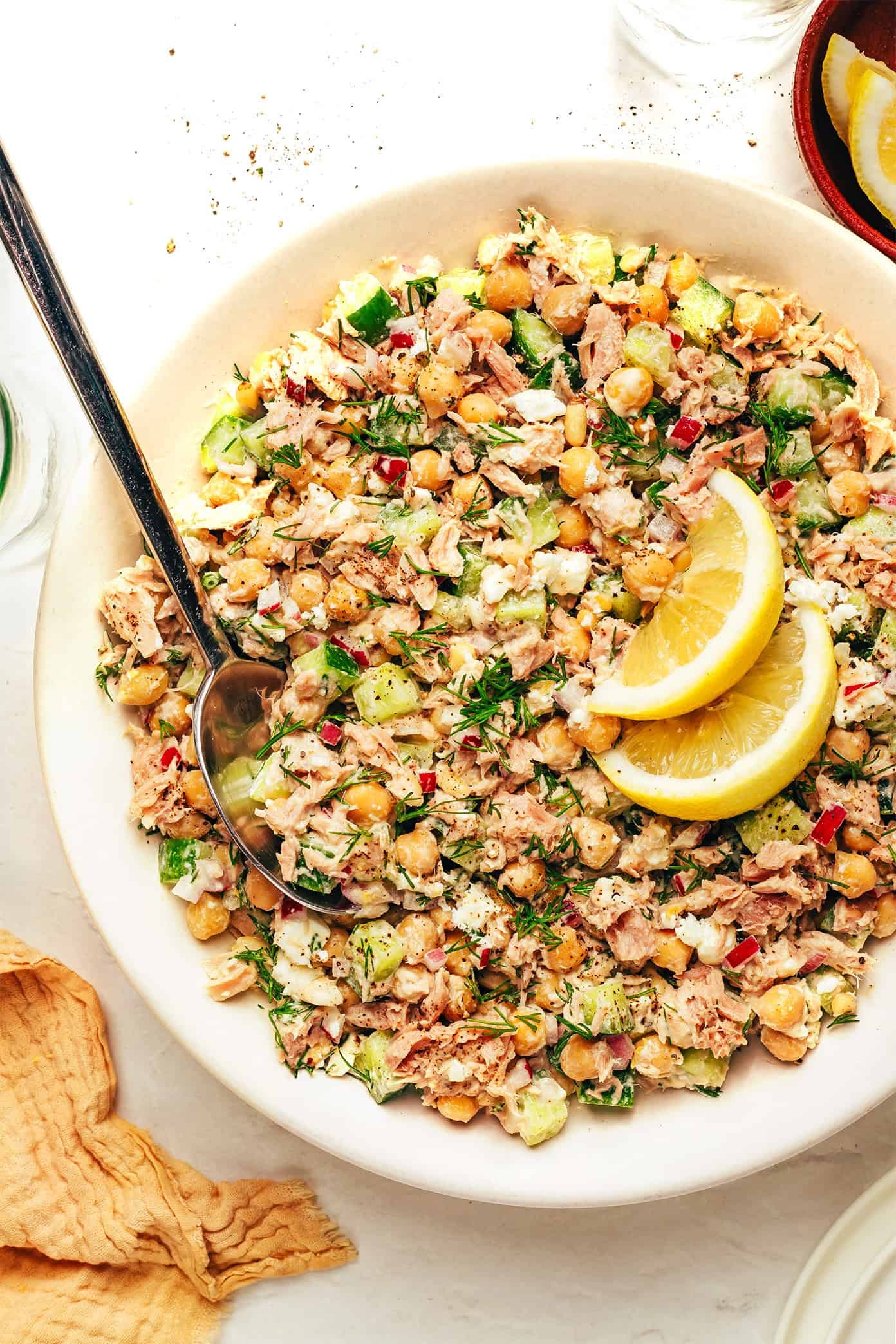 A bowl of tuna chickpea salad with diced vegetables, garnished with two lemon wedges and herbs, with a spoon on the side