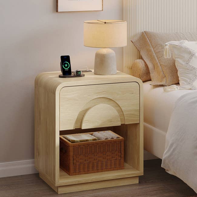 Wooden bedside array  with a lamp, telephone  charging connected  top, and a wicker handbasket  connected  the little   shelf, adjacent  to a furniture  with a textured headboard