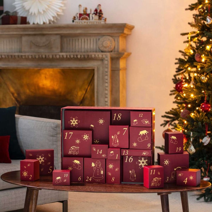 Advent calendar with numbered boxes arranged on a table, set near a Christmas tree and fireplace, suggesting a festive holiday setting