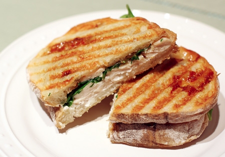 Grilled panini sandwich with melted cheese and arugula on a white plate, cut in half to show filling