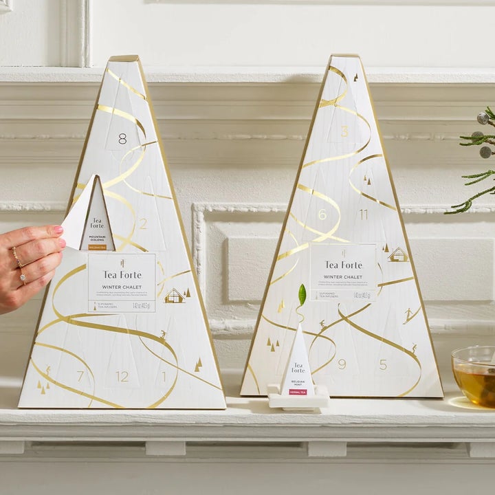 Person opening a slot on a pyramid-shaped advent calendar with tea bags inside, displayed on a shelf next to a teacup