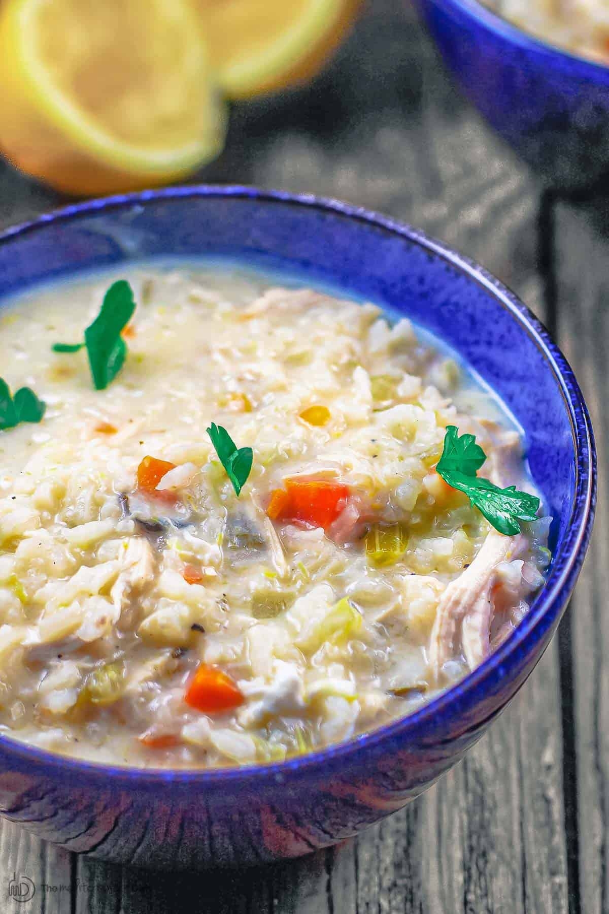 A bowl of creamy chicken and rice soup garnished with parsley, with slices of lemon in the background