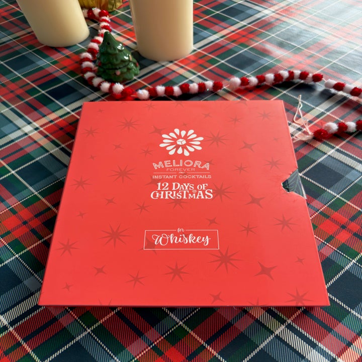 Box of Meliora Instant Cocktails on a plaid tablecloth, labeled "12 Days of Christmas Whiskey." Decorations include candles, a small tree, and garland