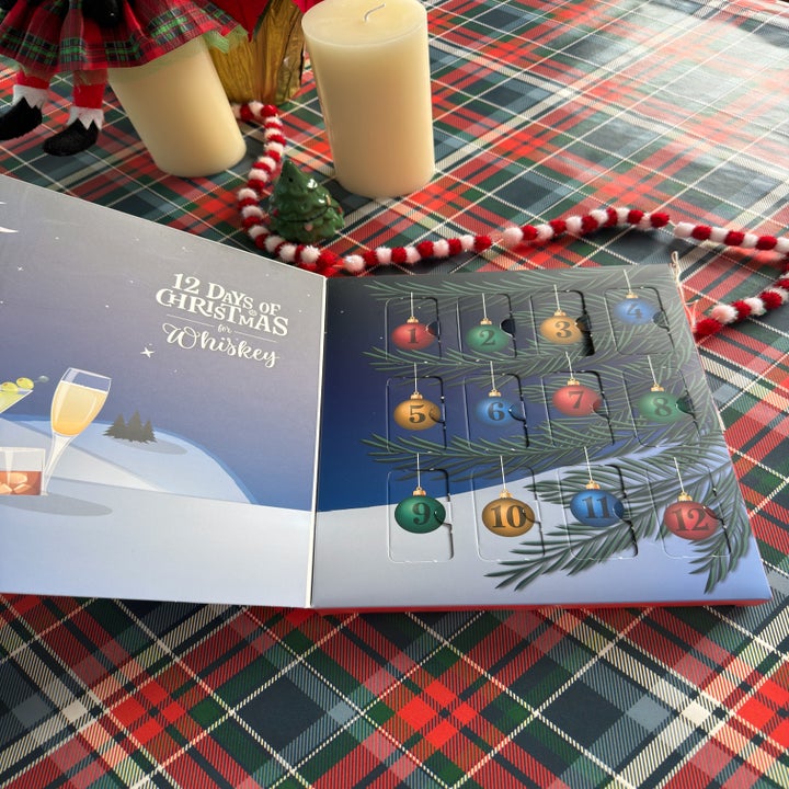 Advent calendar titled "12 Days of Christmas Whiskey" with numbered doors on a holiday-themed tablecloth