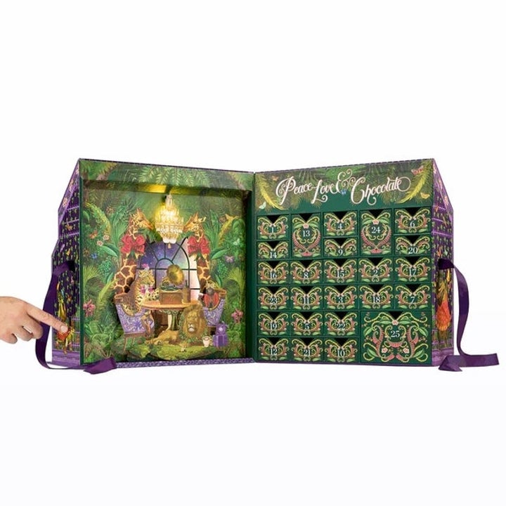 Advent calendar with intricate forest scene, displaying doors labeled with numbers. A hand points to the doors on the right side