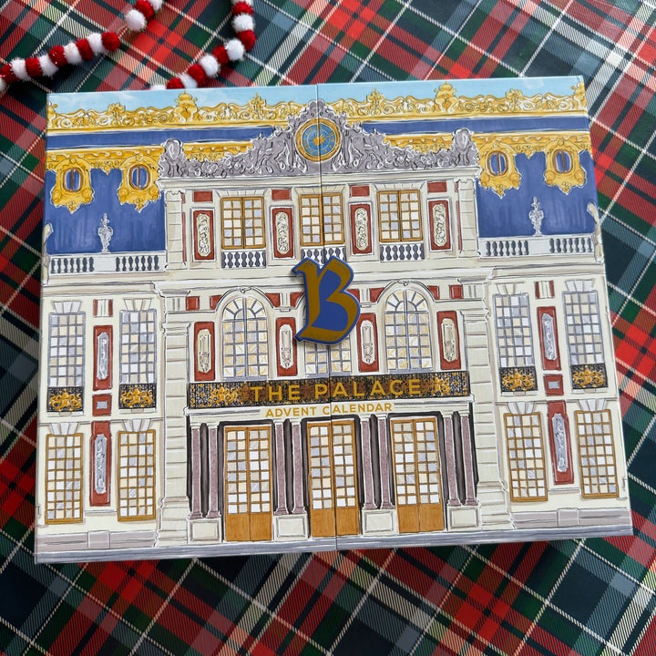 Advent calendar with a palace design and bold "B" on a plaid surface; decorative garland in the background