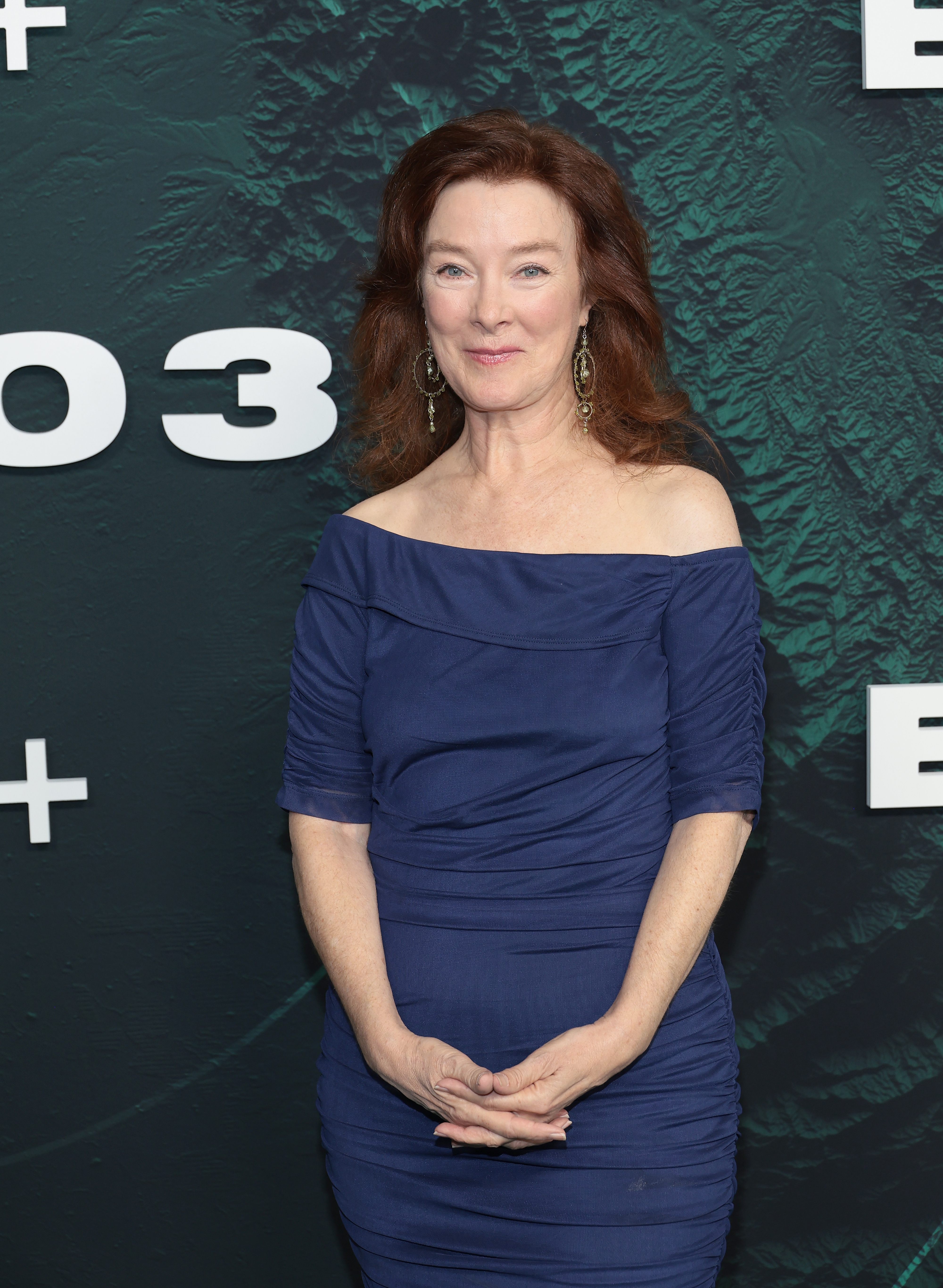 Person på den røde løperen iført en elegant, off-the-skulder blå kjole, stående foran et mønstret bakteppe