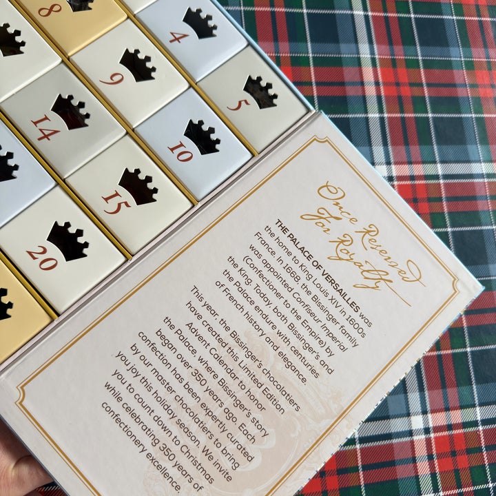 A hand holds a holiday advent calendar with gold and white numbered boxes, featuring text about royal beauty Christmas celebrations