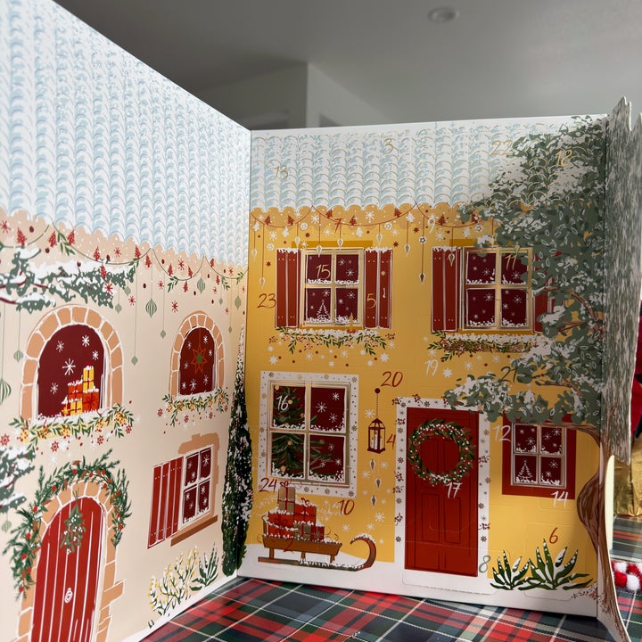 Cozy holiday-themed advent calendar with illustrated house, festive windows, and doors numbered for each day
