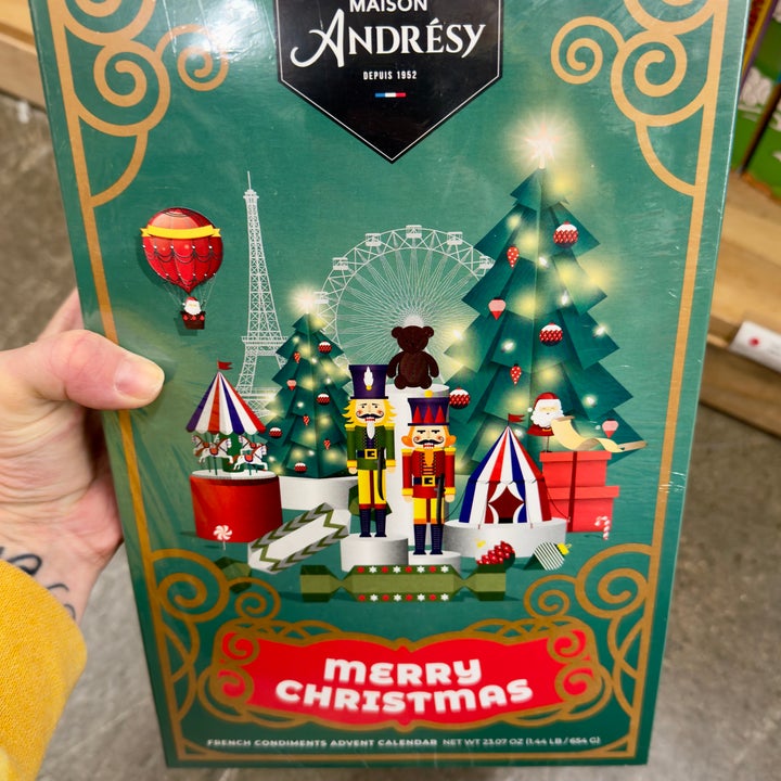 Hand holds an advent calendar featuring nutcrackers, Christmas trees, festive decorations, and the text "Maison Andrésy" and "Merry Christmas."