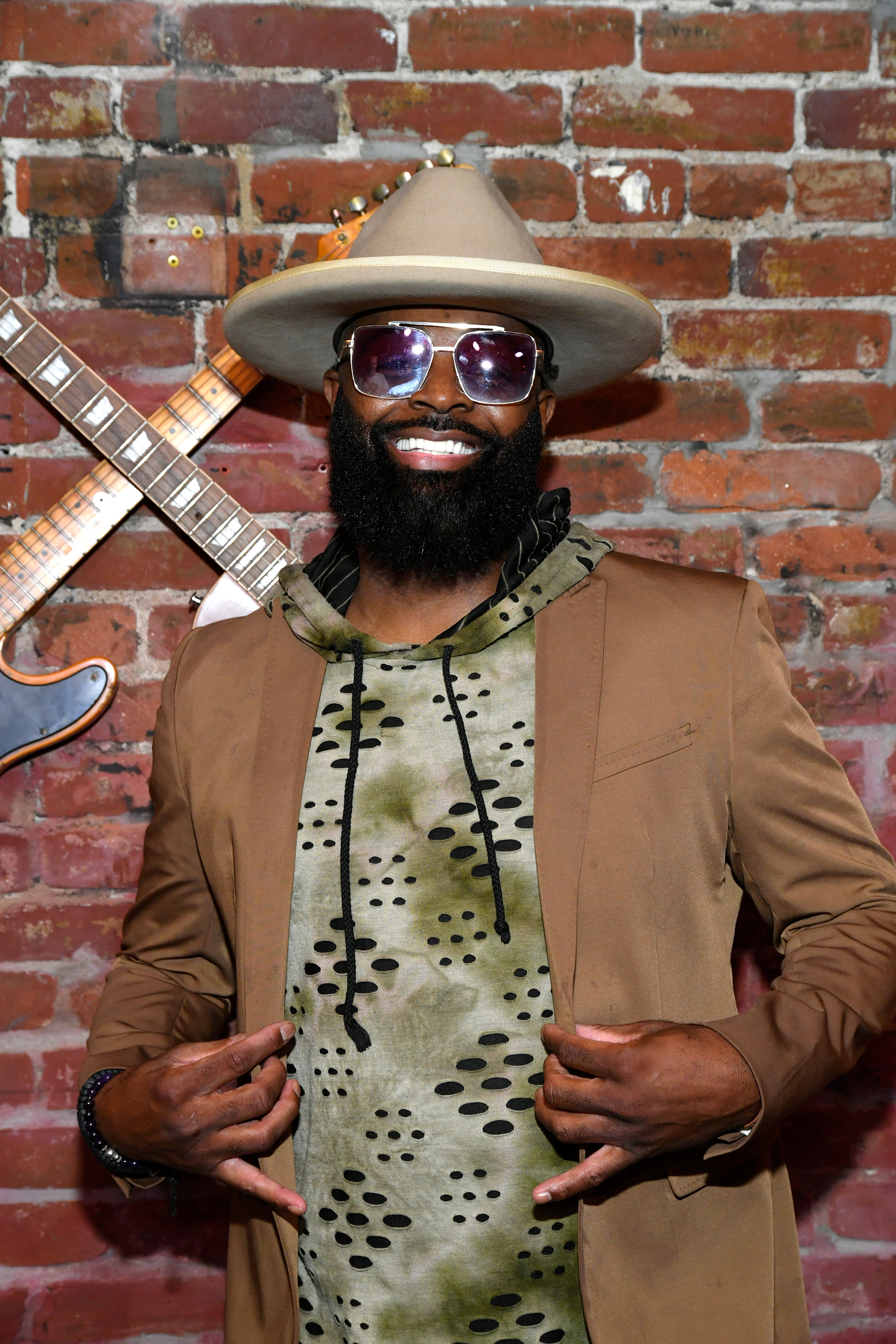 Mann i fedora iført en mønstret hettegenser under en blazer, poserte mot en murvegg med to gitarer