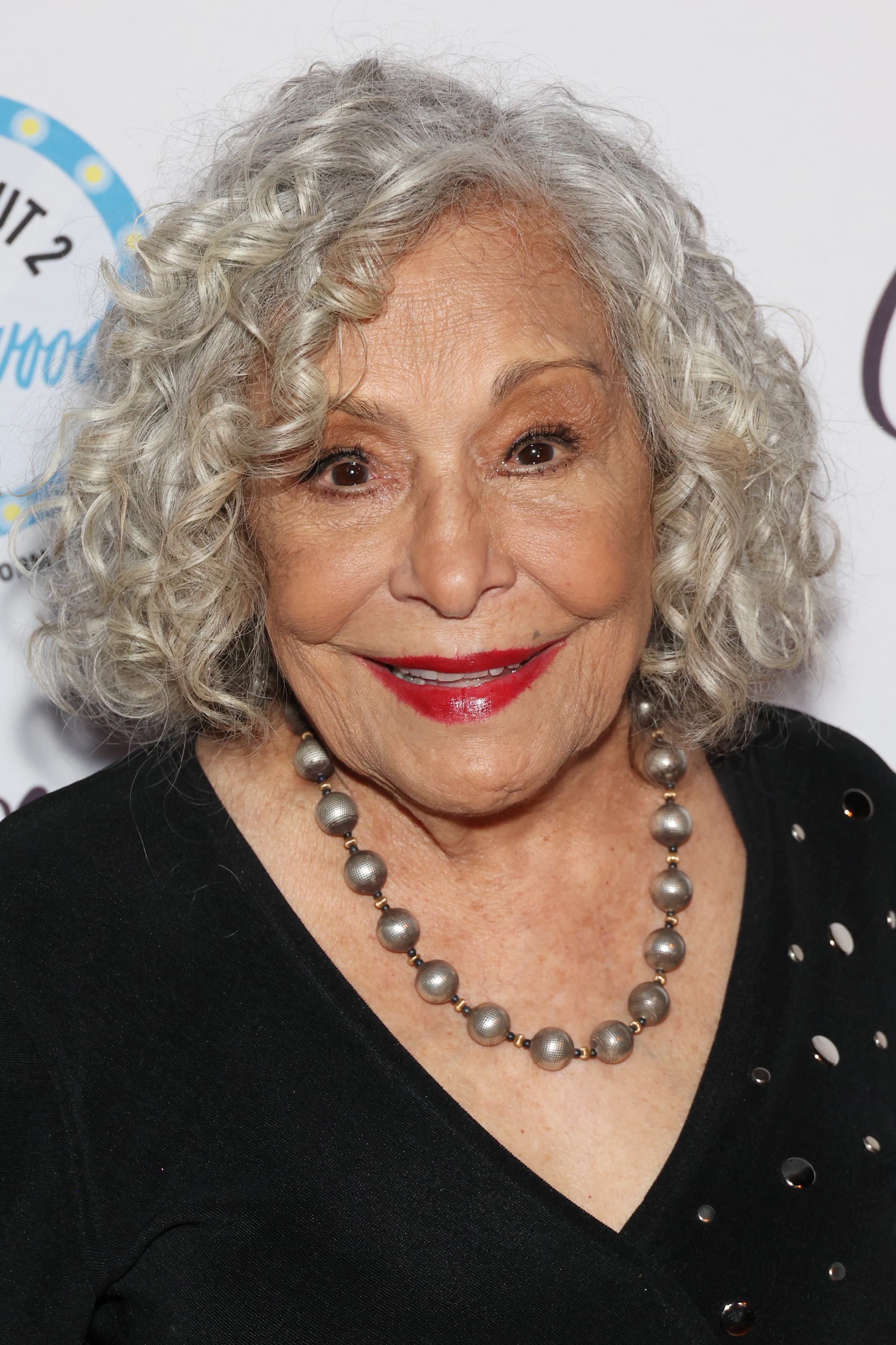 Eldre kvinne med krøllete hår, iført et perlekjede og svart antrekk med dekorative elementer, smiler ved en formell begivenhet