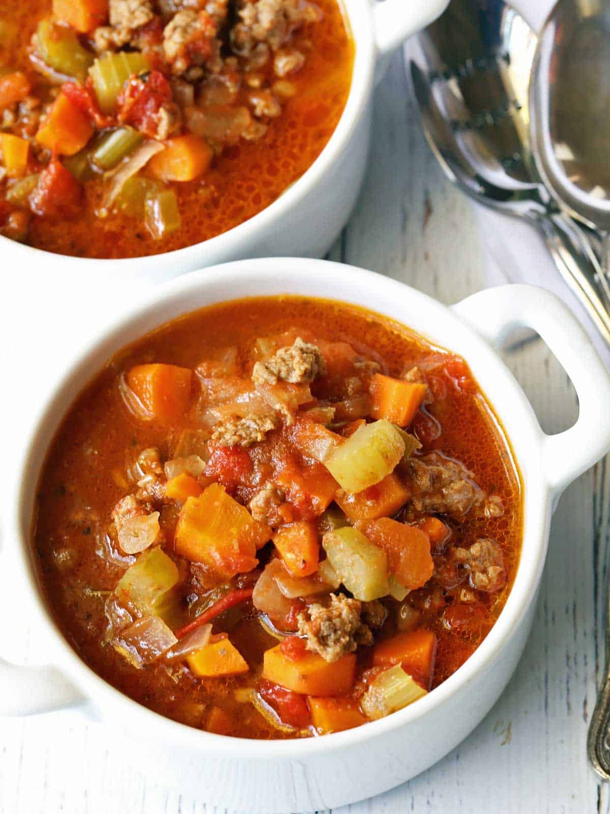 Two bowls of hearty vegetable and beef soup on a table with spoons nearby