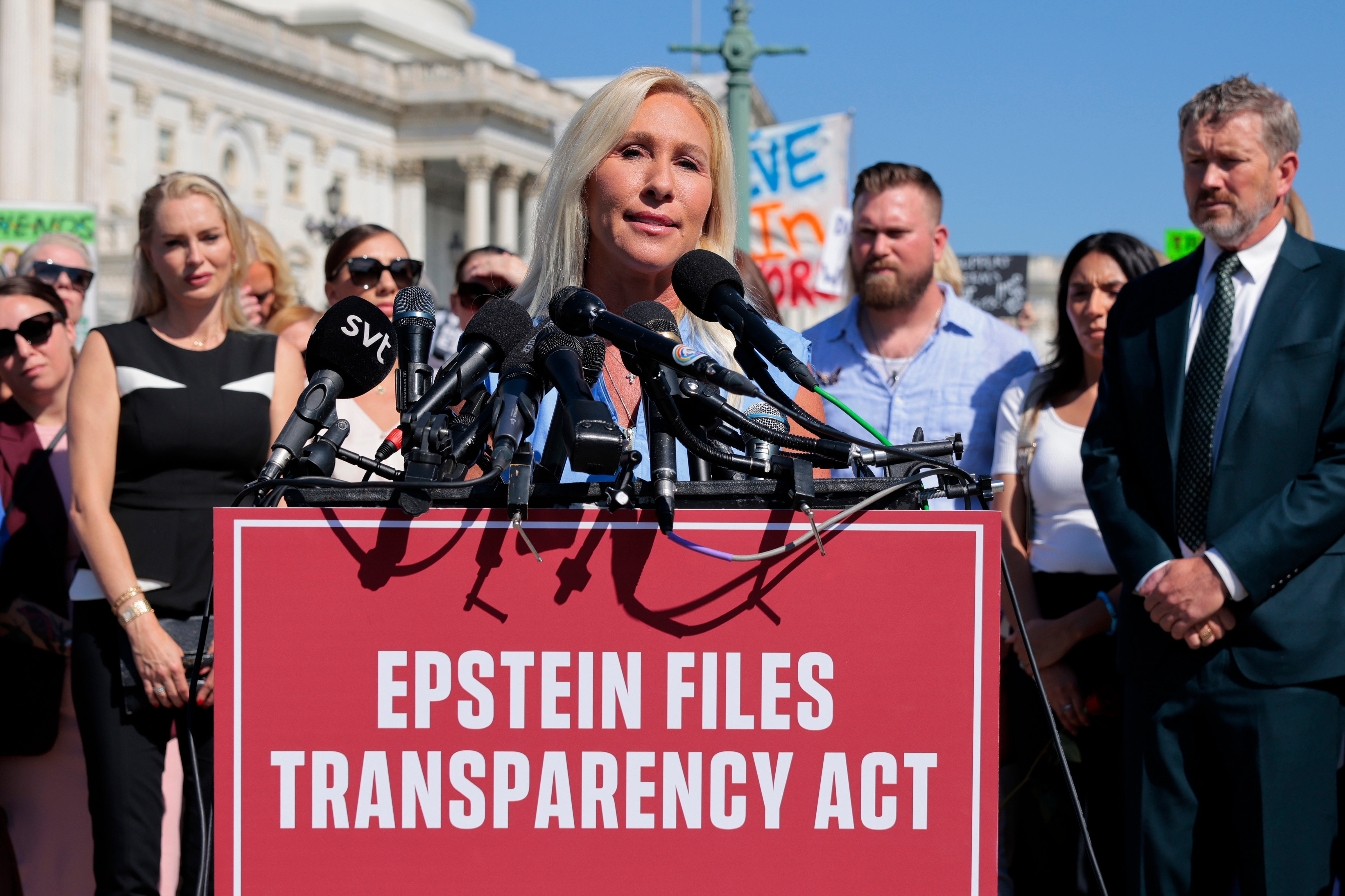 Person speaking at a press event with &quot;Epstein Files Transparency Act&quot; displayed, surrounded by microphones and attendees in business attire