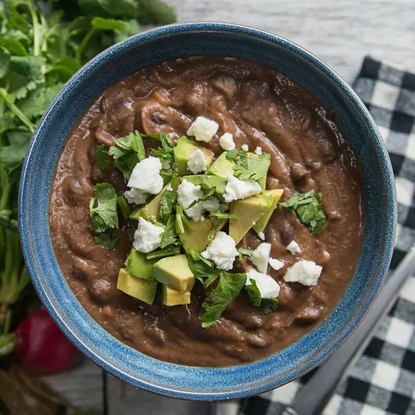 Bowl of black bean soup topped with avocado, feta cheese, and cilantro, next to a checkered napkin and fresh greens