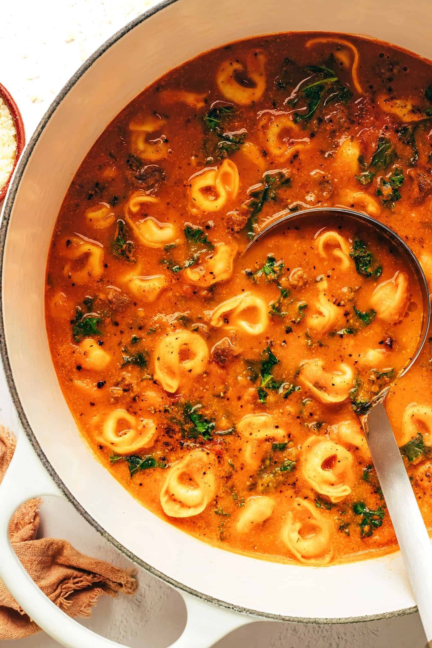 A pot of creamy tortellini soup with spinach and visible chunks of tomato and herbs, with a ladle resting inside
