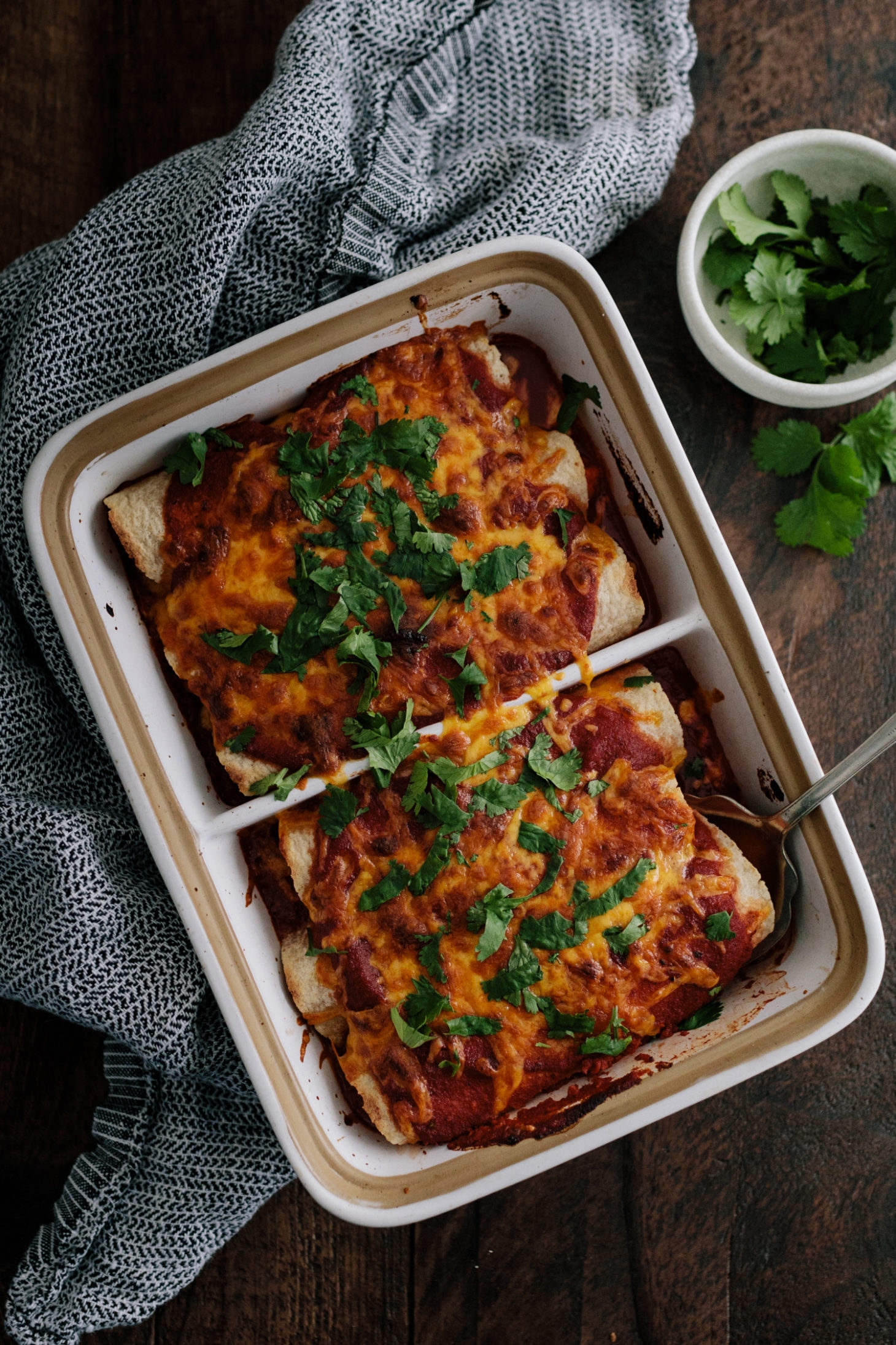 Baked enchiladas topped with melted cheese and cilantro in a dish, with a side of cilantro in a small bowl