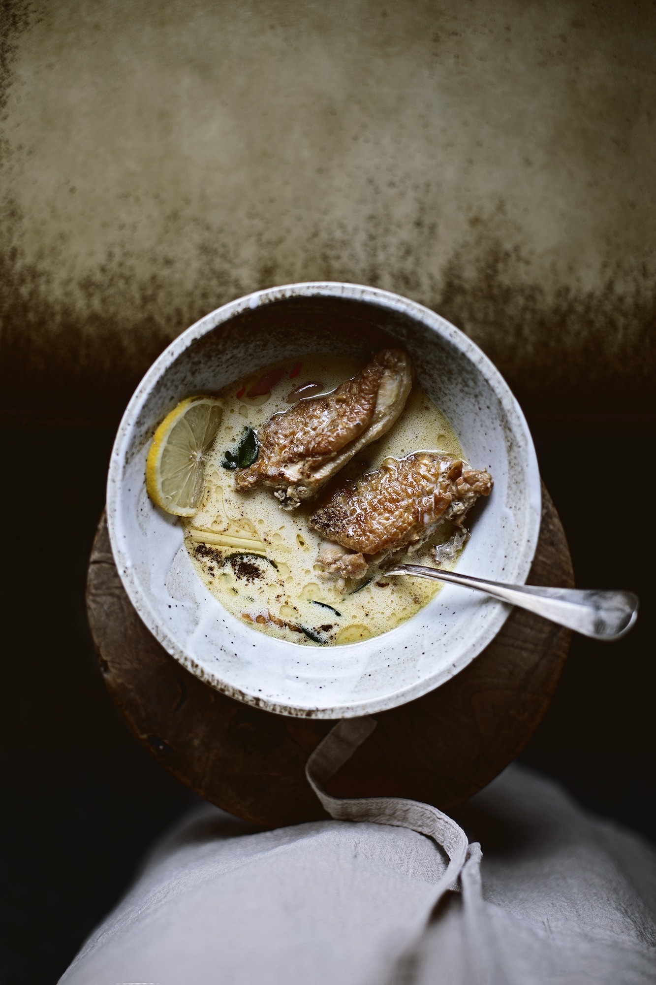 A bowl of creamy soup with a lemon slice, topped with two cooked chicken pieces, on a wooden surface. A spoon rests in the bowl