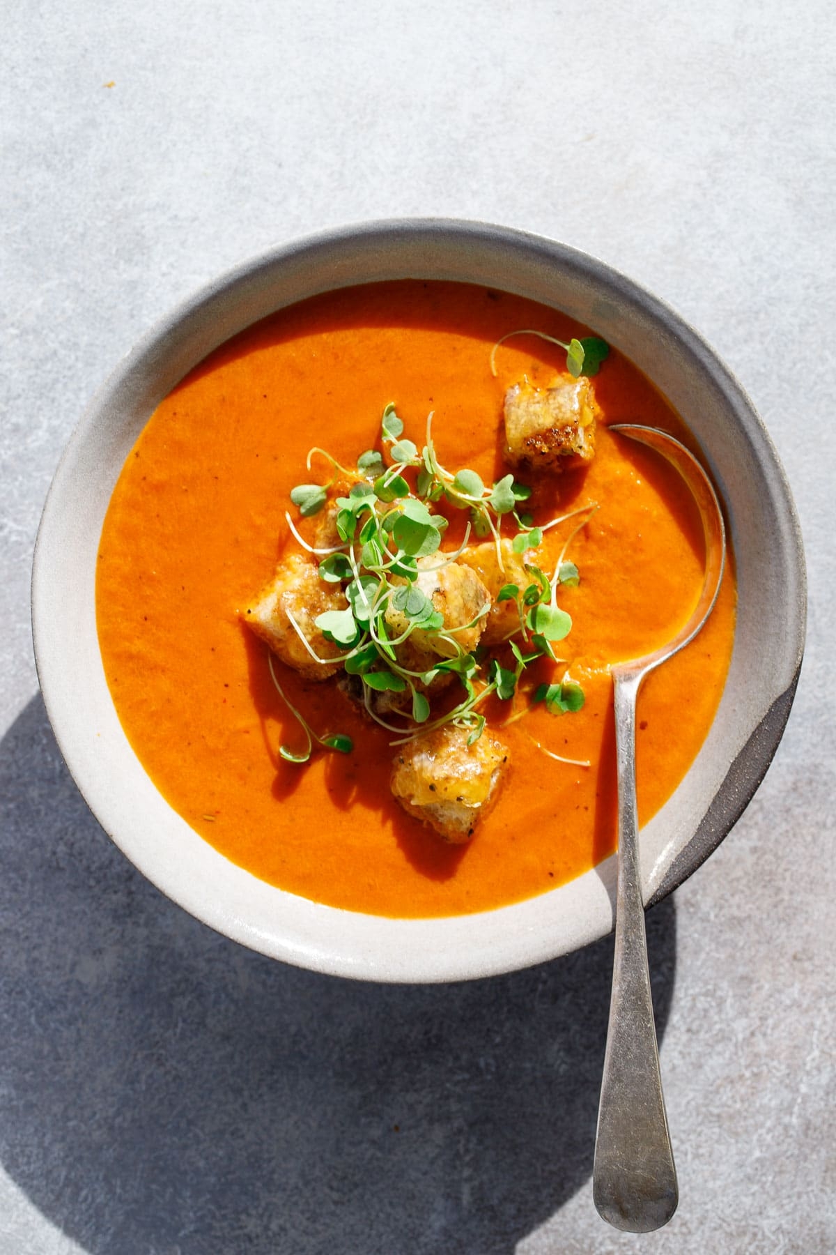 Bowl of creamy tomato soup topped with croutons and fresh herbs, with a spoon resting inside