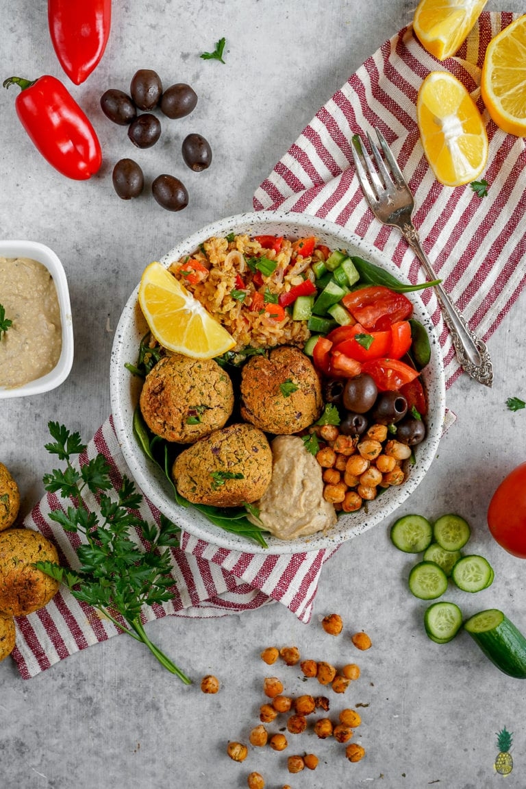 A bowl with falafel, hummus, chickpeas, tomatoes, cucumber, and rice, garnished with parsley and lemon. Surrounded by olives, peppers, and more veggies
