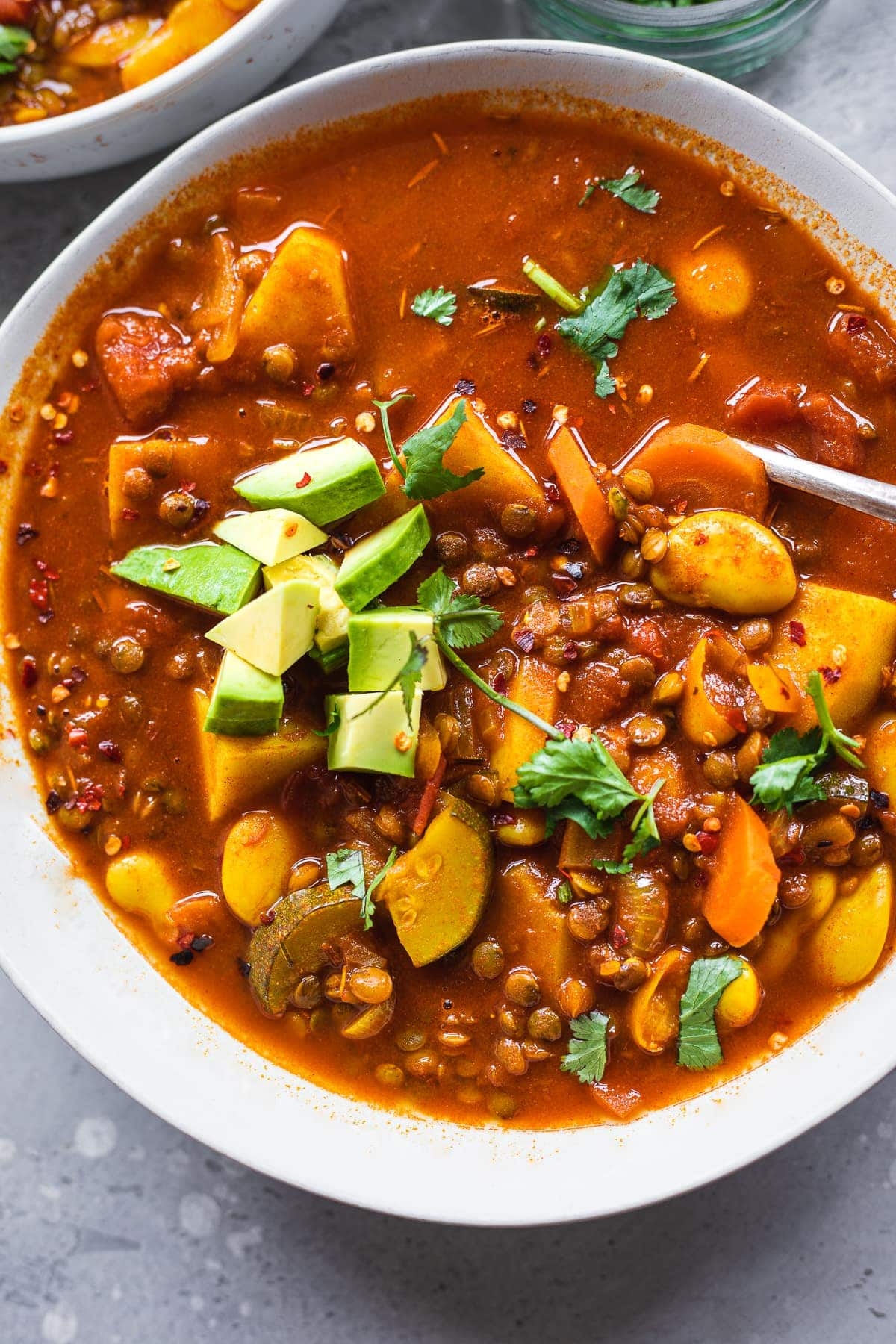 A bowl of hearty vegetable stew with lentils, carrots, zucchini, and avocado chunks, garnished with cilantro