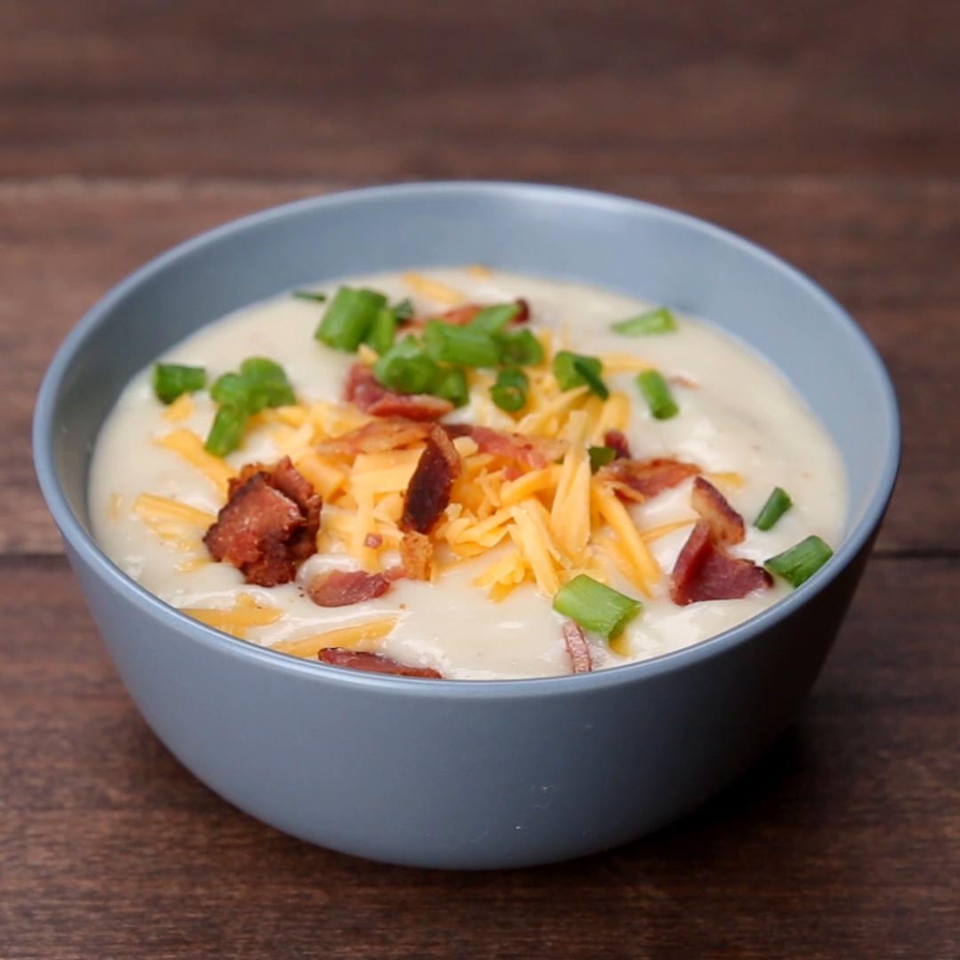 Bowl of creamy soup topped with shredded cheese, bacon bits, and chopped green onions on a wooden surface