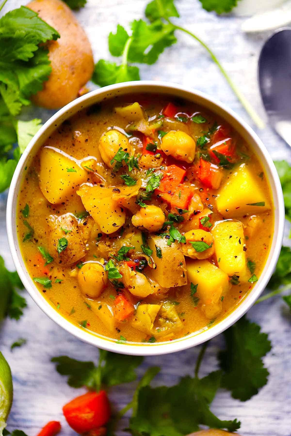Bowl of vegetable and chickpea soup garnished with herbs, surrounded by fresh ingredients like cilantro, potato, and bell pepper on a wooden surface