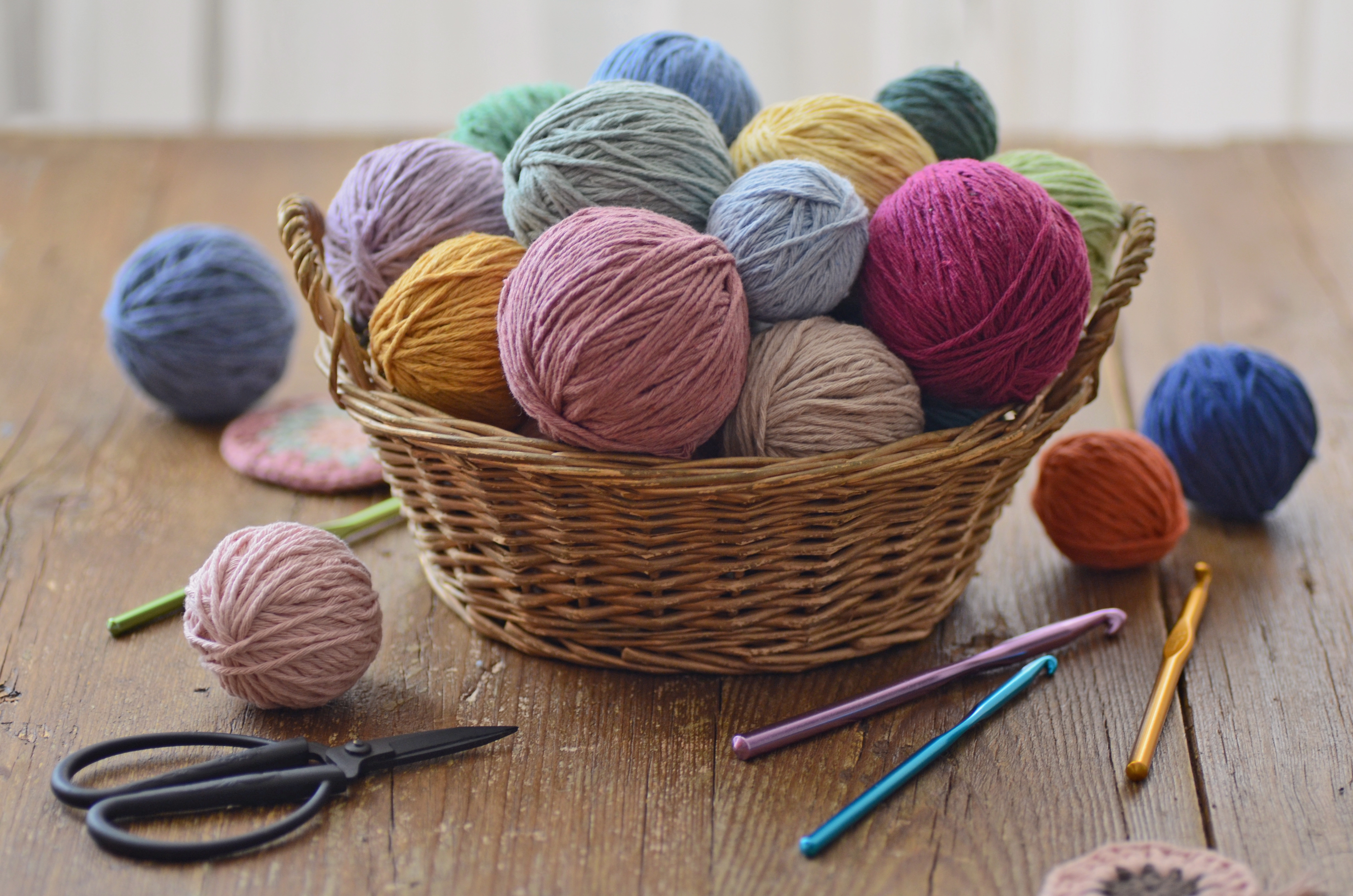 Wicker handbasket filled with balls of yarn connected a woody table, surrounded by crochet hooks and scissors