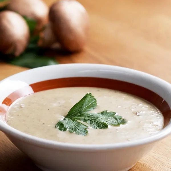 Bowl of creamy mushroom soup garnished with parsley, with whole mushrooms in the background on a wooden surface
