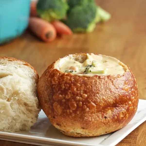 Soup served in a bread bowl on a plate, with broccoli and carrots in the background