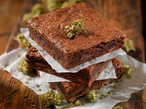 Three chocolate brownies stacked on parchment paper, surrounded by scattered cannabis buds on a wooden surface