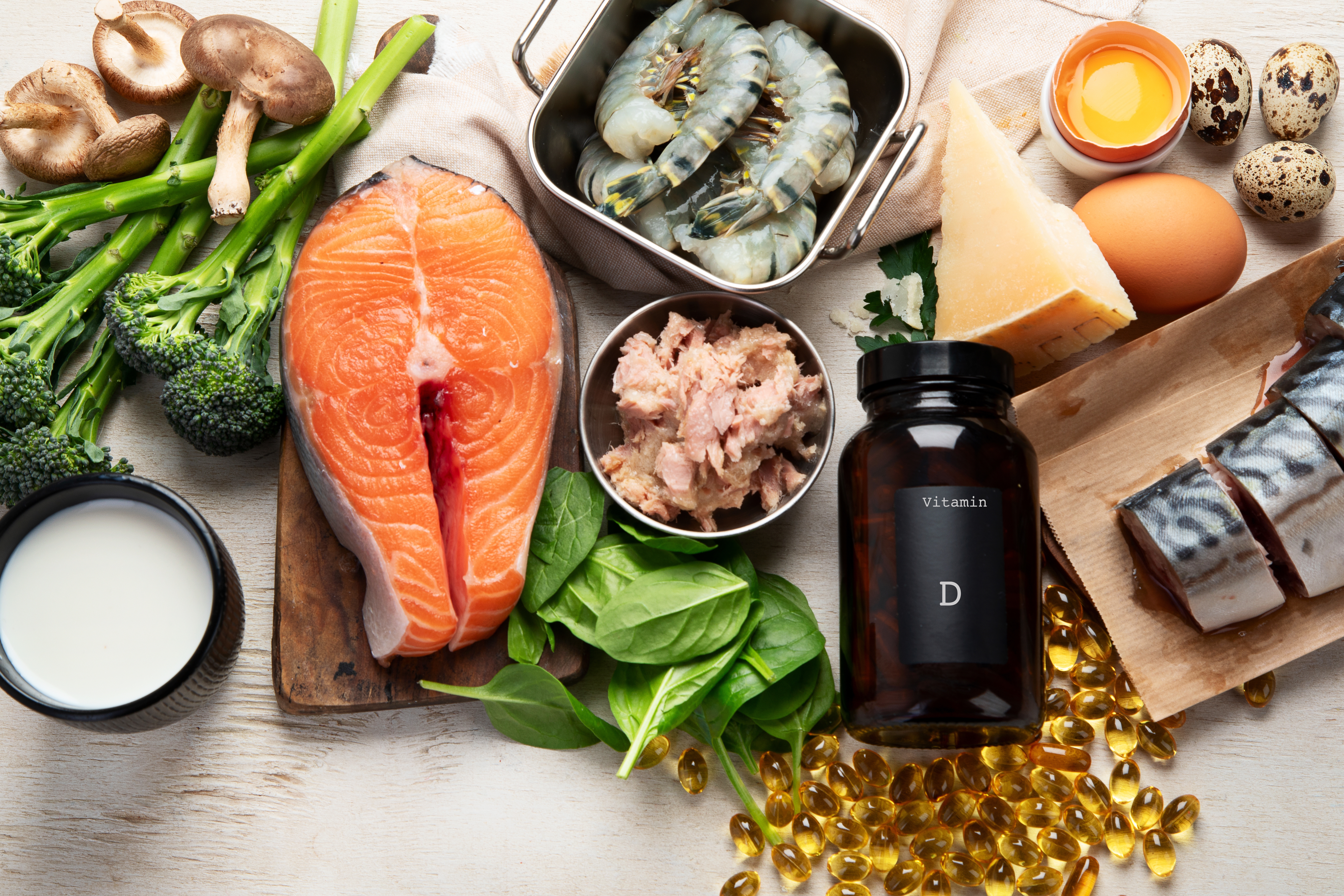 Assorted vitamin D-rich foods including salmon, tuna, ovum yolk, mushrooms, spinach, cheese, shrimp, broccoli, milk, and supplements connected a table