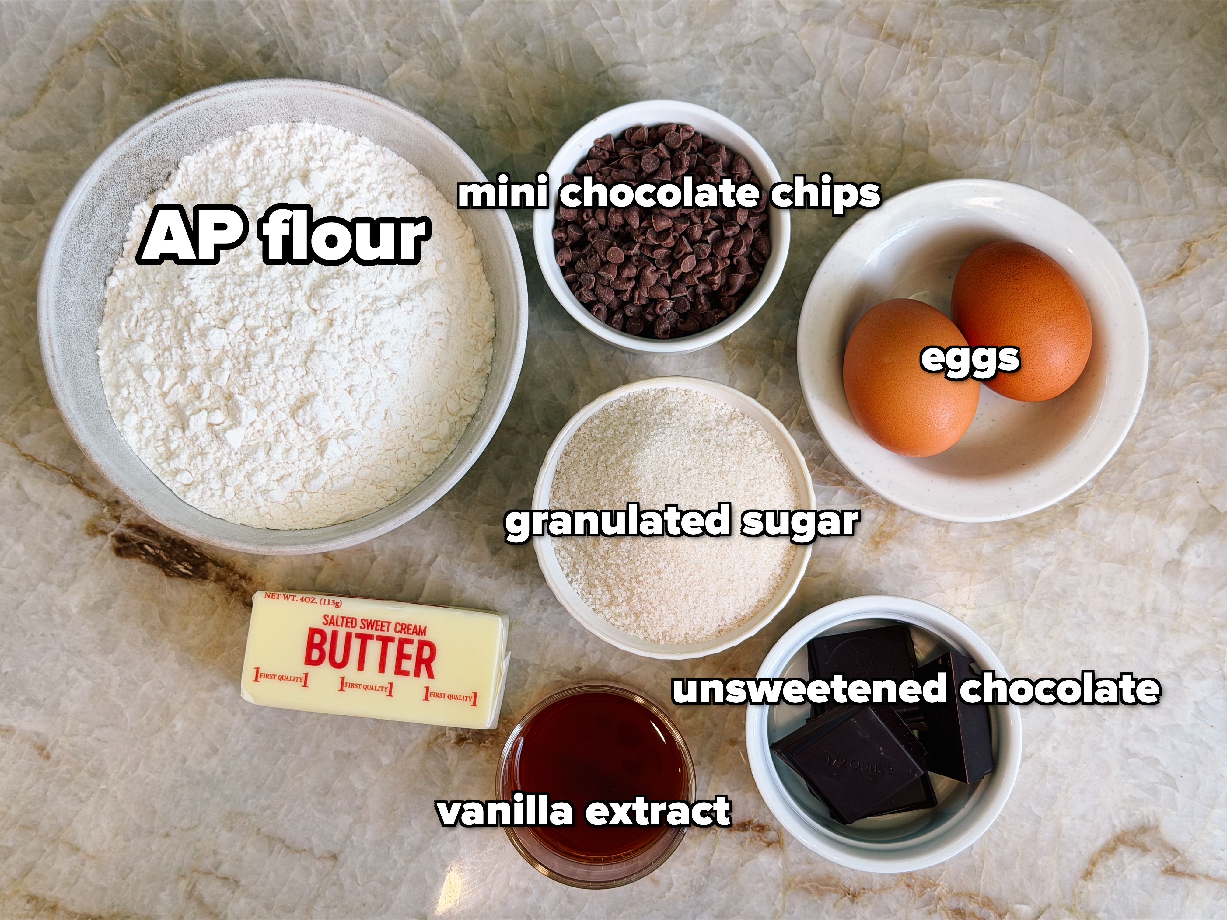 Baking ingredients on a counter: flour, chocolate chips, eggs, butter, granulated sugar, dark chocolate, and vanilla extract