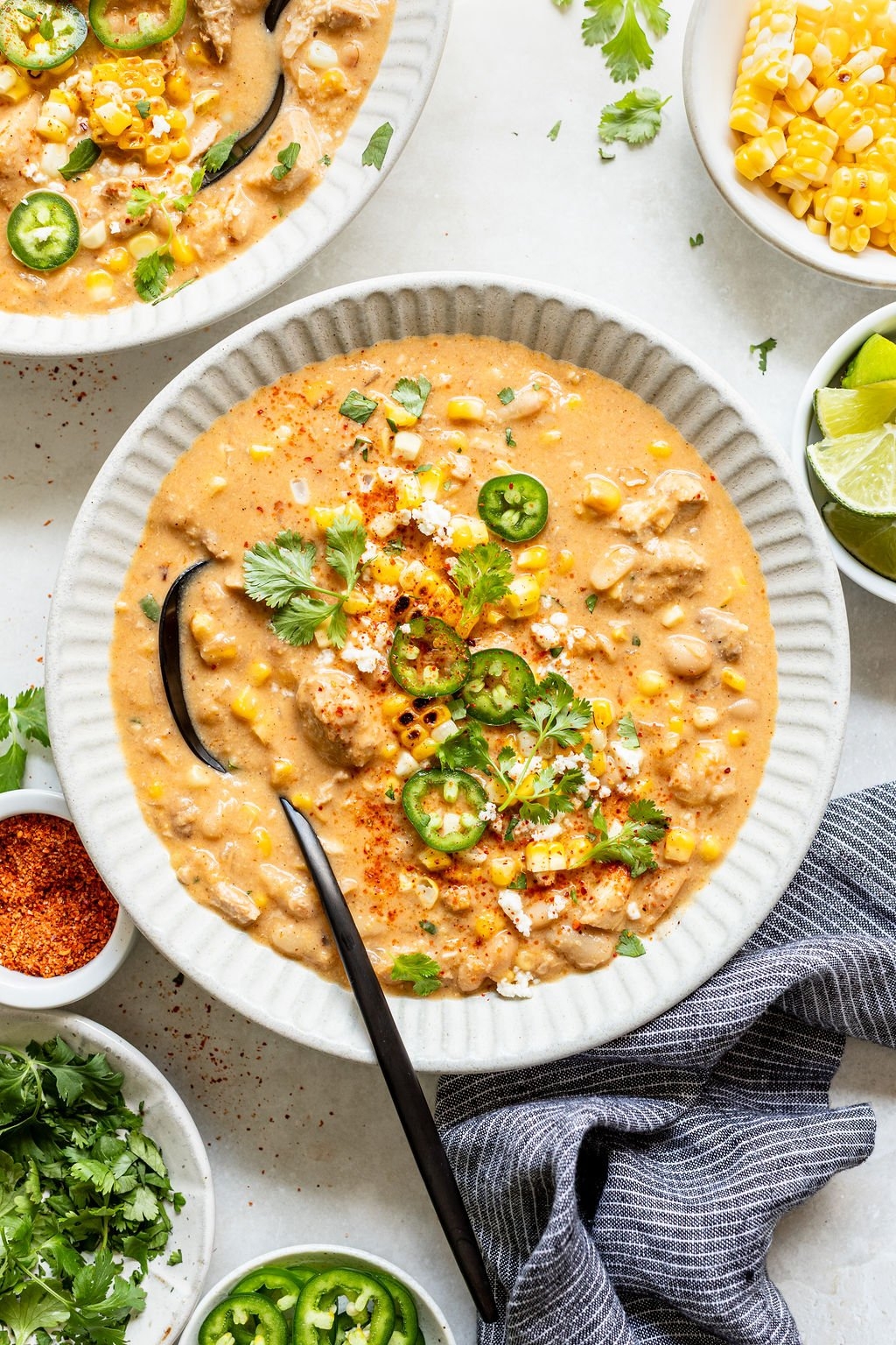 Bowl of creamy corn and chicken soup garnished with sliced jalapeños, cilantro, and cheese, surrounded by small dishes of spices and lime wedges