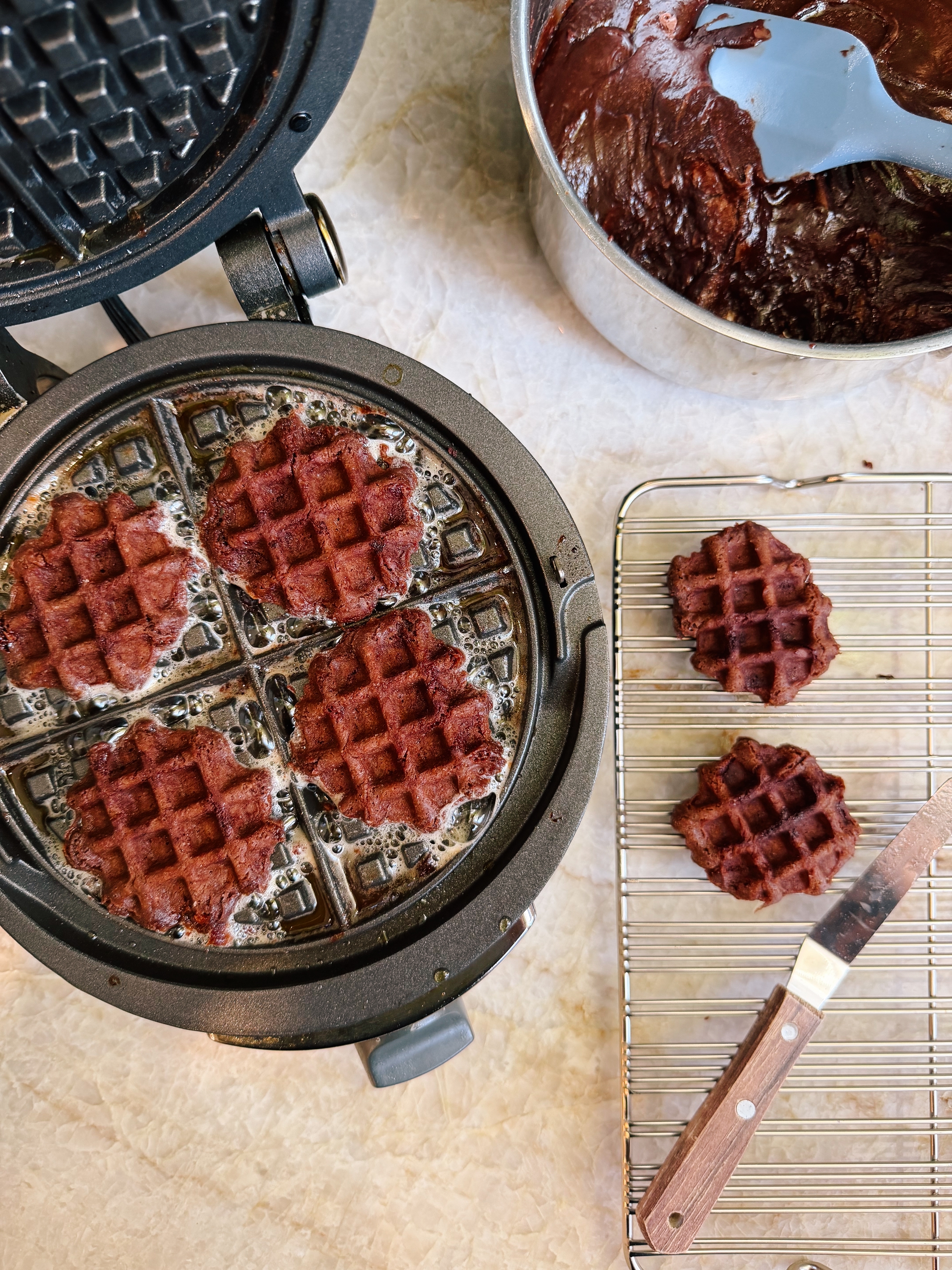 Open waffle maker with five chocolate waffles. A bowl with waffle batter is beside it, and two waffles are cooling on a rack with a knife nearby