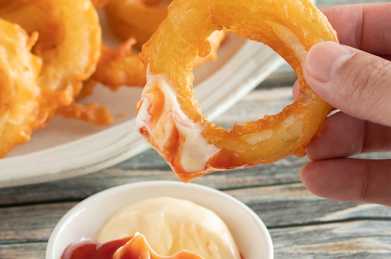 手が玉ねぎリングを持ち、ケチャップとマヨネーズが入った小皿にディップしています。
