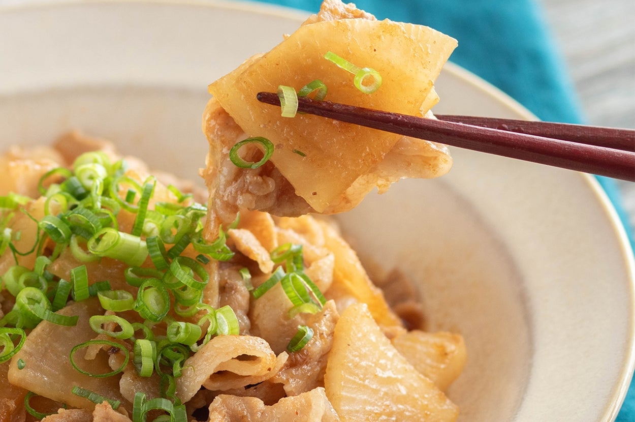 薄切り豚肉と大根の煮物が白い皿に盛られ、箸で持ち上げられている。刻みネギがトッピングされている。