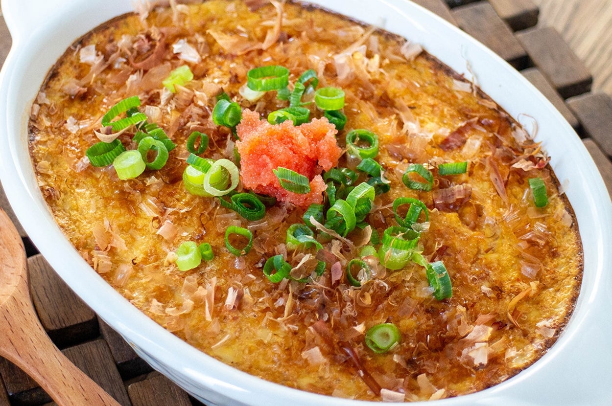 オーブン焼きされたディッシュ（緑の薬味と明太子トッピング）、木製マットの上に白い皿で盛りつけられている。