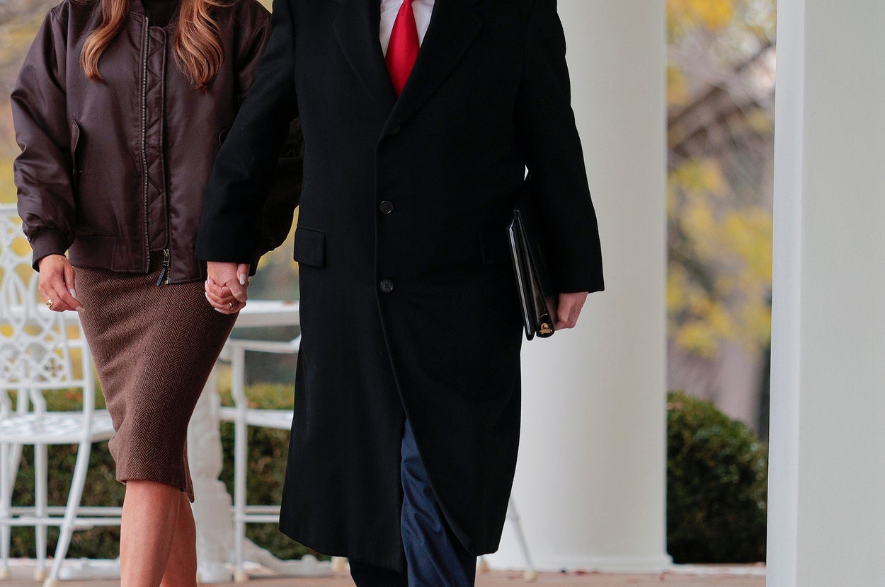男女が庭を歩く。女性はジャケットとスカートを着用し、男性は黒いコートに赤いネクタイを着けている。