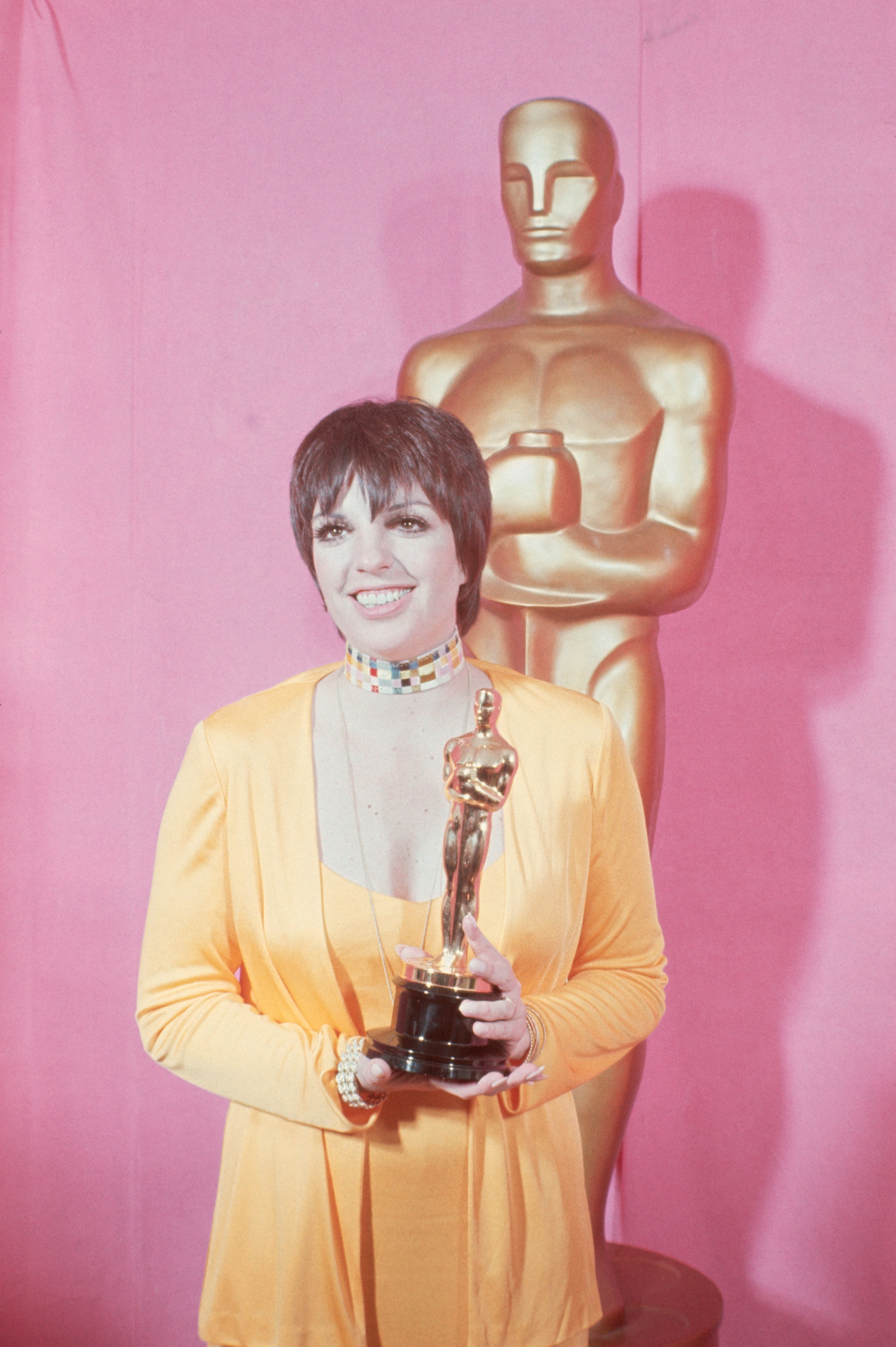 Person holding an Oscar statuette, wearing a V-neck formal  and a beaded choker, lasting  successful  beforehand   of a ample  Oscar statue
