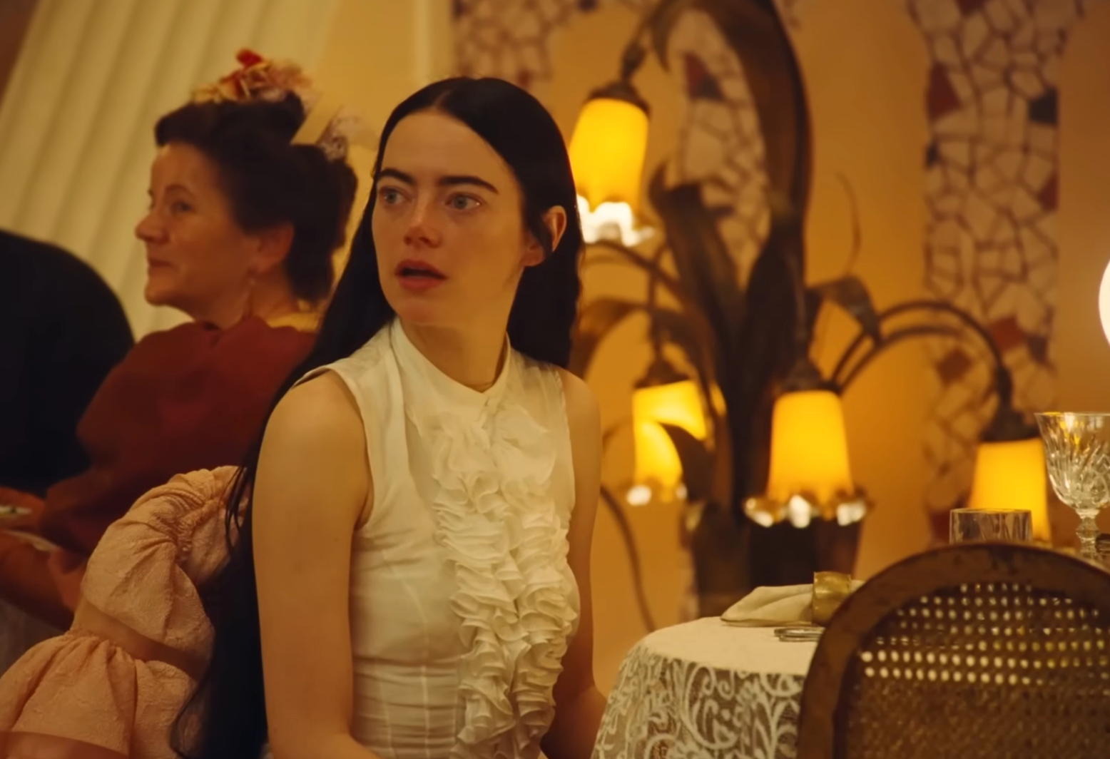 A idiosyncratic   with agelong  hairsbreadth  wearing a ruffled achromatic  blouse sits astatine  a eating  table, looking to the broadside  successful  a dimly lit, ornate room