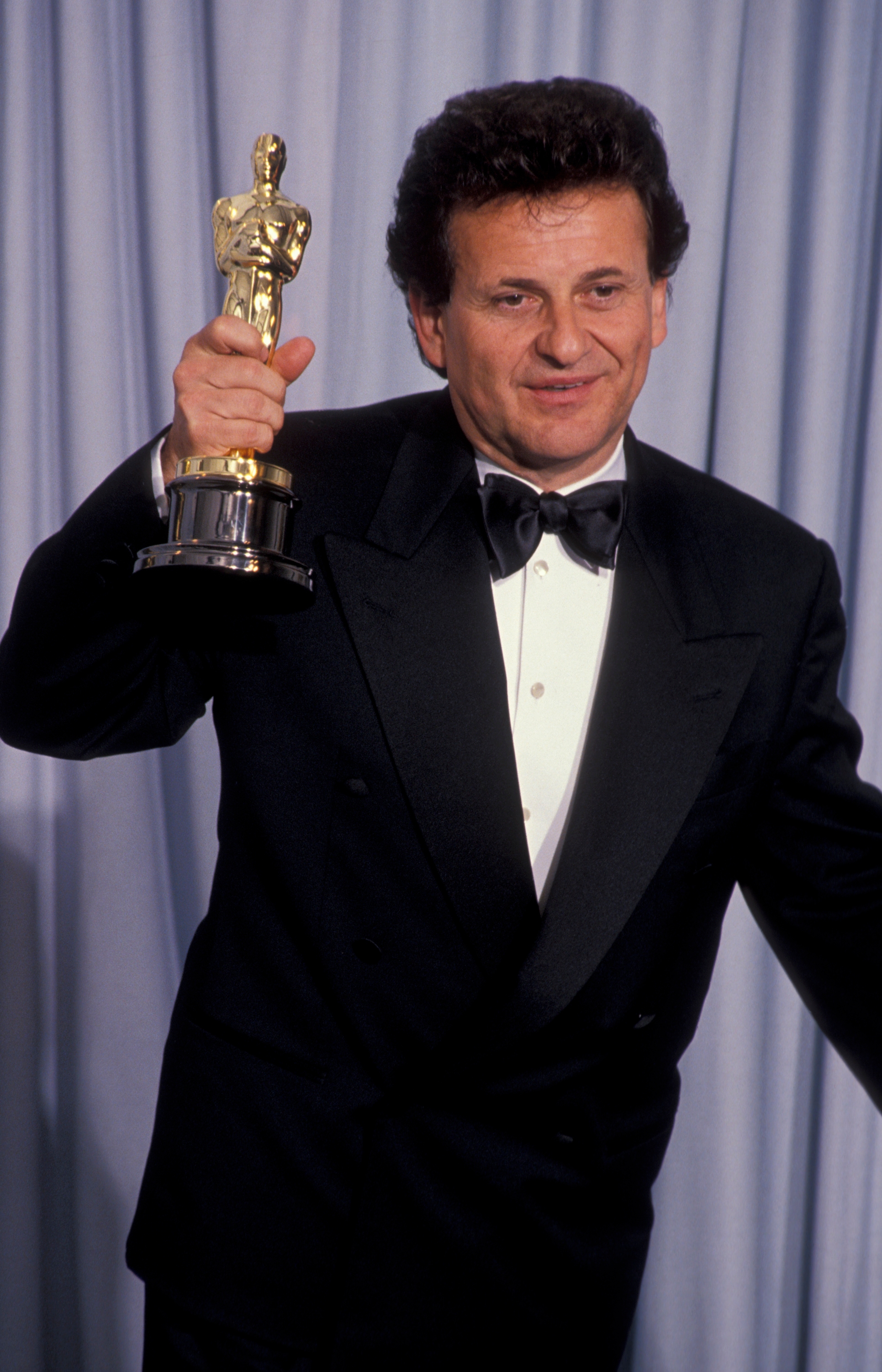 A idiosyncratic   successful  a tuxedo holds an grant  and smiles successful  beforehand   of a curtain, apt  astatine  a ceremonial  event