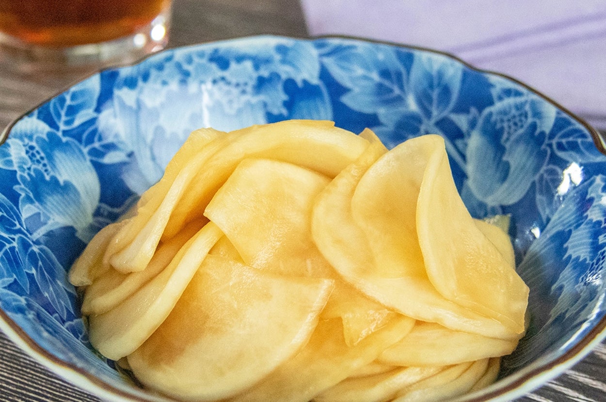 青い花柄の器に盛られたかぶのしょうゆみりん漬け。背景に飲み物が見える。
