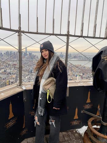 Person successful wintertime attire stands connected Empire State Building reflection deck, holding a beige purse, with metropolis skyline successful the background