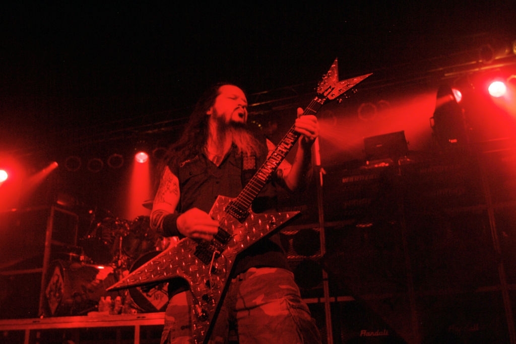 Musician playing guitar with intensity on stage, surrounded by vibrant stage lights, wearing a sleeveless vest and pants