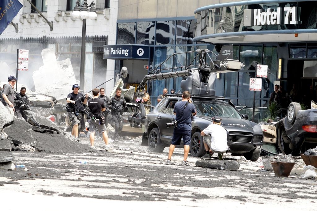 Movie scene being filmed in urban setting with crashed cars and debris, crew managing camera equipment around a car, Hotel 71 in background