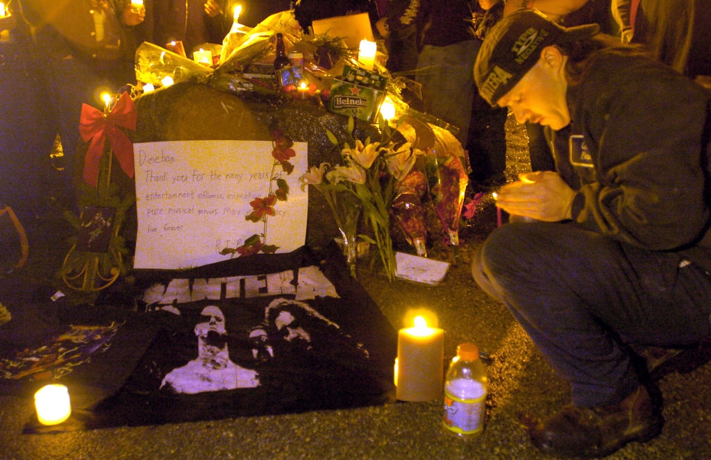 Person kneels at a candle-lit vigil with a &quot;Pantera&quot; shirt and flowers, honoring someone
