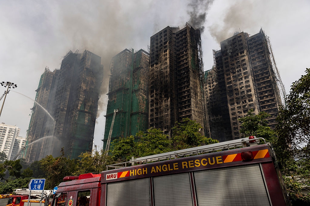 Smoke billows from high-rise buildings during a fire, with a high angle rescue vehicle in the foreground. Firefighters manage the scene